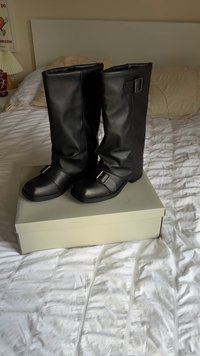 Bottes noires montantes en simili cuir, présentant un bout arrondi, un tige large et un accent en boucle argentée au niveau de la cheville. Présentées sur un fond clair.