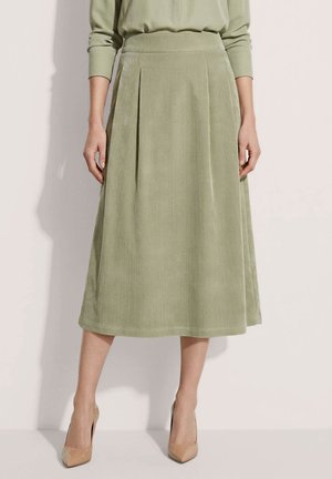 Femme portant une jupe midi en velours côtelé vert clair avec des plis, associée à un haut assorti et des talons hauts beiges pointus.
