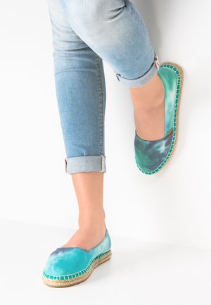 Espadrilles in blauw-groene tie-dye met een ronde neus, canvas bovenwerk en jute zool. Rolled denim jeans complementeren de schoenen.