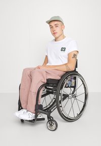 Una persona en una silla de ruedas lleva una camiseta blanca con gráficos verdes y pantalones rosa claro. Tiene una gorra gris y zapatillas blancas.