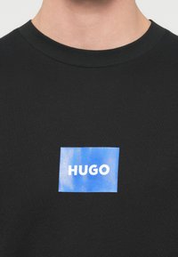 Juoda medvilninė marškinėliai su mėlynu stačiakampiu lipduku centre, kuriame baltais rašmenimis parašyta „HUGO“. Apvalus kaklo iškirptė ir trumpomis rankovėmis.