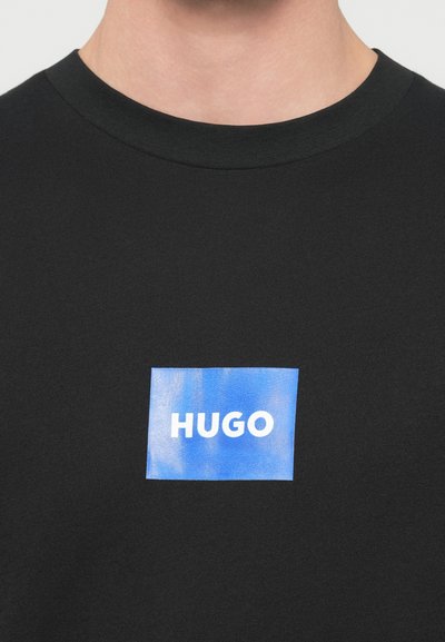 Черна памучна тениска с квадратен син етикет и бял надпис "HUGO" в центъра. С кръгло деколте и къси ръкави.