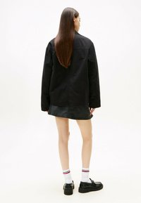 Chaqueta de camisa de algodón negra con un corte relajado, combinada con una falda de mezclilla. El atuendo incluye zapatos negros y calcetas blancas con rayas rojas.