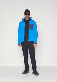 Mėlyna fliso striukė su užtrauktuko kišene ir juodais akcentais, derinama su juodomis kelnėmis ir sportbačiais. Modelis dėvi akinius nuo saulės ir kepurę.