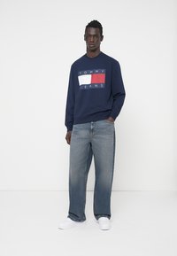 Felpa blu navy con logo bianco e rosso, vestibilità rilassata. Abbinata a jeans in denim azzurro e sneakers bianche, in piedi contro uno sfondo neutro.
