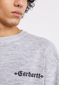 Grå striktrøje med rund hals, tekstureret stof og sort "Carhartt" logo på venstre bryst.