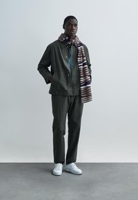 Olivovo zelená bunda a nohavice, doplnené vzorovaným viacfarebným šálom. Biele tenisky dokončujú outfit. Hladká látka, voľný strih.