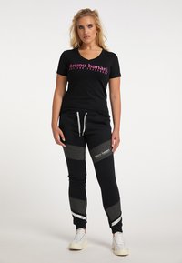 Schwarzes V-Ausschnitt-T-Shirt mit pinkem Logotext, kombiniert mit schwarzen Jogginghosen mit grauen Akzenten und weißen Streifen an den Bündchen. Weiße Sneaker.