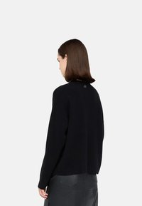 Maglione nero lavorato a maglia con vestibilità rilassata, dettagliato con un piccolo logo sul retro del collo. Indossato con una gonna in pelle nera.