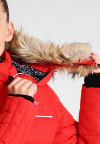 Chaqueta acolchada roja con capucha forrada de piel, con un forro interior estampado y bolsillo con cremallera. Tela texturizada y puños elásticos visibles.