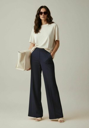 Femme portant un t-shirt crème oversize, un pantalon large bleu marine, des talons ouverts beige, des lunettes de soleil foncées, tenant un grand sac fourre-tout crème.