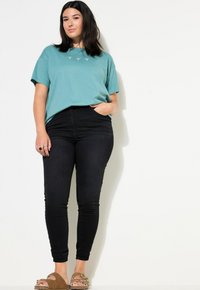 Camiseta de algodón verde azulado de manga corta con bordado en el frontal, combinada con jeans negros ajustados y sandalias marrones con una correa de acento.