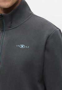 Grå fleecejakke med høy krage, med blå glidelås og en liten logo med "1962" på brystet. Myk tekstur og avslappet design.