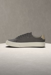 Baskets basses grises avec semelle blanche et languette de talon beige, présentées sur une surface claire avec un fond beige uni.