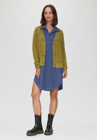 Olivgrüner Strickcardigan mit Kabelmuster, Knopfverschluss, kombiniert mit einem blauen Hemdkleid und schwarzen Stiefeletten mit dicker Sohle.