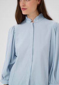 Blouse bleu clair avec un col à volants, des manches ballons et une fermeture à boutons sur le devant. Texture lisse avec un léger éclat et coupe ajustée.