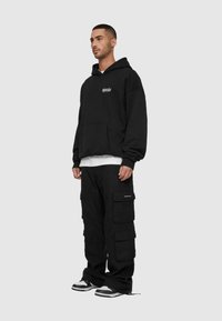 Schwarzer Hoodie mit Fronttasche, lockere Passform. Kombiniert mit schwarzen Cargo-Hosen mit mehreren Taschen. Weißes T-Shirt mit kurzen Ärmeln ist darunter sichtbar.