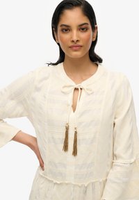 Helle cremefarbene Bluse mit strukturiertem Muster, Schlüsselaugen-Ausschnitt und Bindedetail. Verfügt über weit ausgestellte Ärmel und goldene Quastenakzente.