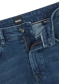 Light blue denim jeans with a zip fly and button closure, featuring a leather brand patch and contrast stitching along the waistband.