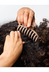 Beige Plastik-Haarklammer mit breiten Zähnen, die dunkles, lockiges Haar hält; eine Hand positioniert die Klammer, während die andere das Haar stützt.