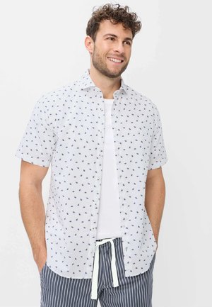 Mann mit lockigem Haar, der ein weißes Kurzarmhemd mit kleinen blauen Mustern über einem weißen T-Shirt und gestreiften Hosen trägt.