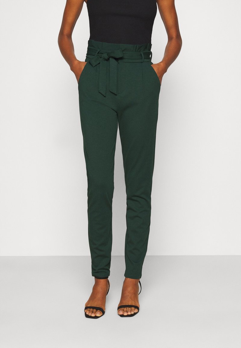 Pantalon vert taille haute avec ceinture à nouer, coupe slim, poches latérales et texture de tissu lisse, associé à des talons noirs à lanières.