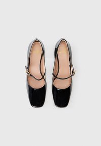 Sapatos Mary Jane de verniz preto com bico quadrado, uma tira fina no peito do pé e um detalhe de fivela dourada. No interior, apresenta um logótipo em forma de coração.