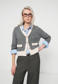 Gray and beige cardigan featuring a v-neck, long sleeves, two front pockets, and buttons. Worn over a blue striped shirt and gray trousers.