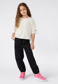 Top corto de color beige claro, pantalones cargo negros con bolsillos laterales y zapatillas rosas con malla texturizada y suelas blancas. Acentos del logo de Champion.