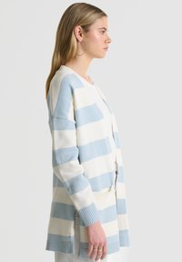 Lengvas kardiganas su plačiomis mėlynomis ir baltomis juostomis, raišteliais rankovėse, atviru priekiu ir dviem priekinių kišenių. Minkšta megztinė tekstūra.