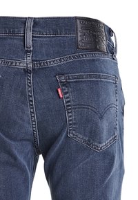 Jean denim bleu foncé à coupe classique, avec un patch de marque en cuir à la taille et des coutures distinctives sur les poches arrière.