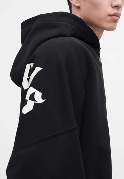 Jeune personne portant un sweat à capuche noir avec un motif abstrait blanc sur l'épaule, vue de profil sur un fond uni.