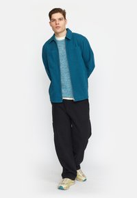 Turquoise zip-up jack over een gestructureerde blauwe trui, gecombineerd met losse zwarte broek en beige sneakers met blauwe accenten. Twee borstzakken zichtbaar.