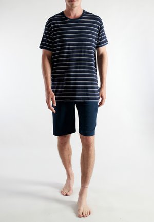 Man standing barefoot wearing navy blue shorts and a navy blue t-shirt with white horizontal stripes against a plain white background.