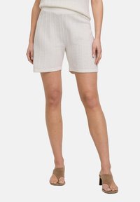 Shorts blancs en tricot avec une texture côtelée verticale, dotés d'une ceinture large et de revers roulés, associés à des talons à enfiler marron.