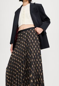 Zwarte plisseerok met een gouden en witte paisleypatroon, gecombineerd met een cropped crèmekleurig topje en een zwarte blazer. Soepele textuur, op maat gemaakt.