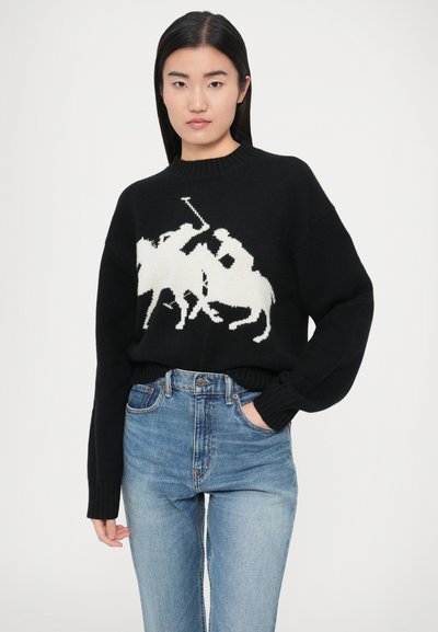 Črni pleteni pulover z belo konjeniško grafiko, rebrastim izrezom okrogle oblike in dolgimi rokavi, v kombinaciji s svetlo modrimi kavbojkami z visokim pasom.