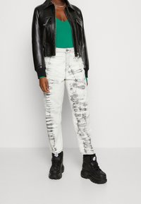 Schwarze Leder-Cropped-Jacke mit zwei Fronttaschen, kombiniert mit weißen Jeans mit schwarzen Batik-Mustern und schwarzen klobigen Sneakers.