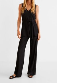 Schwarzes ärmelloses V-Ausschnitt-Top mit einem gegürtelten Bund und locker sitzenden schwarzen Hosen. Das Outfit wird mit flachen schwarzen Sandalen kombiniert.