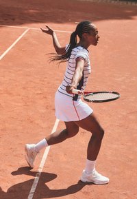 Jugadora de tenis con el pelo trenzado, vistiendo una falda blanca y una camiseta a rayas, preparándose para golpear la bola en una pista de tierra batida.