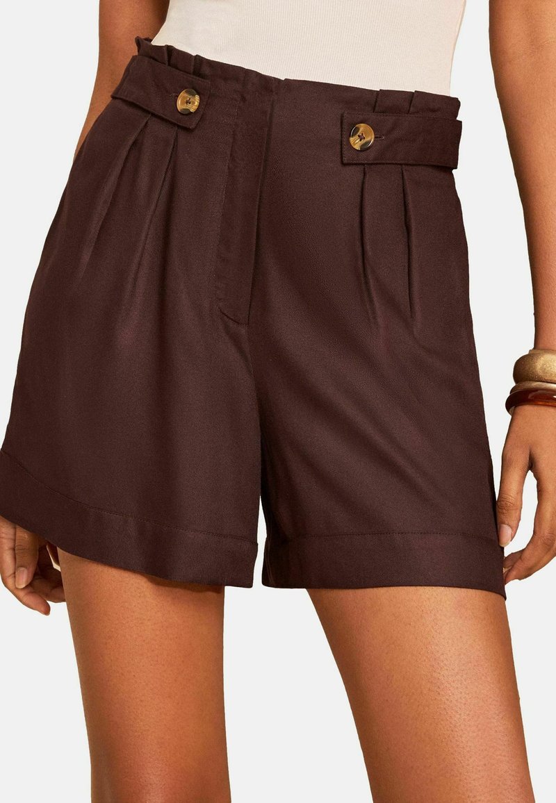 Frau trägt dunkelbraune Shorts mit hoher Taille und Falten, taillierte Knopfleisten, umgeschlagene Säume und ein beiges Oberteil, accessoriert mit Armbändern.