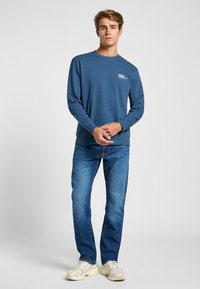 Camisa azul de manga larga con cuello redondo, con un gráfico blanco en el lado izquierdo del pecho, combinada con jeans de mezclilla azul claro y zapatillas beige.