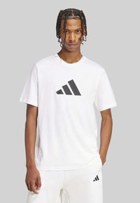 Camiseta blanca de manga corta con un gran logo negro de Adidas en el frente. Hecha de tela suave, con cuello redondo y un corte relajado.