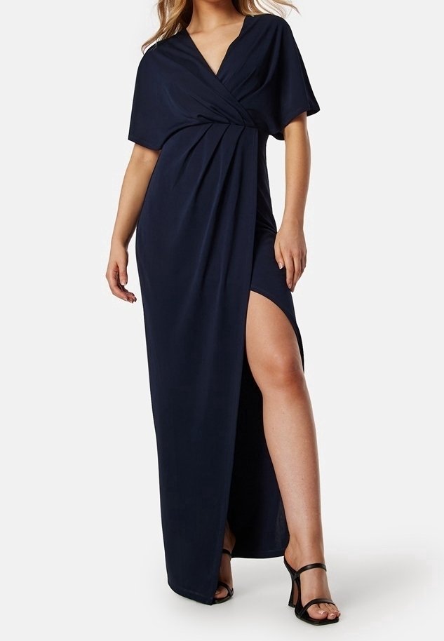 Femme portant une robe portefeuille bleu marine avec une fente haute et des sandales à talons noires, debout devant un fond uni.