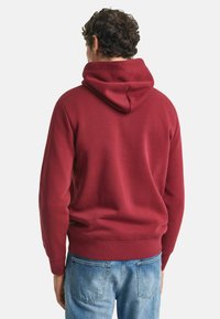 Rode hoodie met een grote verstelbare kap, ribgebreide boorden en zoom. Gemaakt van zachte stof met een gladde textuur. G gedragen met lichtblauwe spijkerbroek.