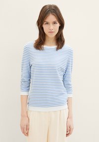 Pull rayé bleu clair en tissu doux avec des manches longues et un bord blanc au niveau du col et des poignets. Associé à un pantalon beige.