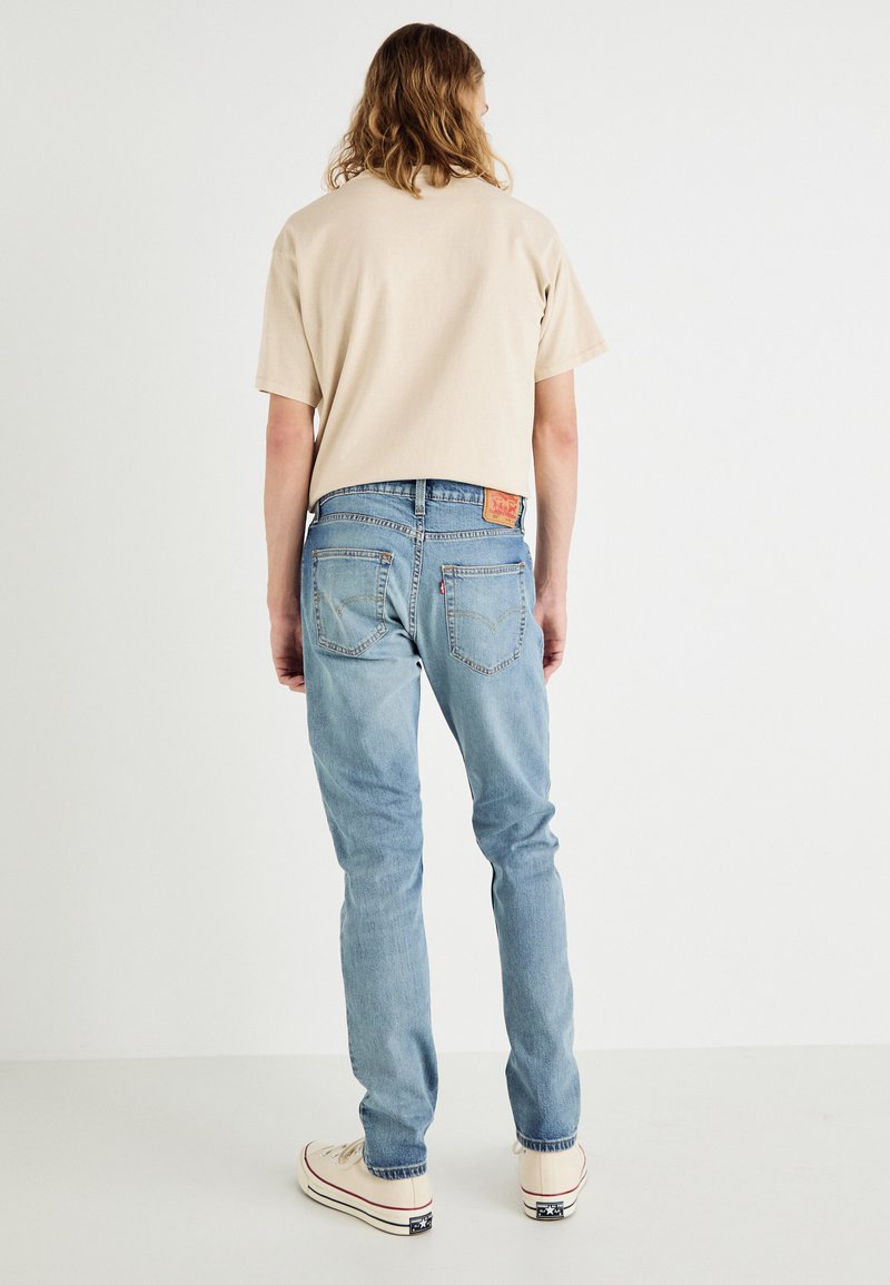 T-shirt en coton beige clair à manches courtes, associé à un jean en denim bleu clair avec poches arrière et un patch logo à la taille.