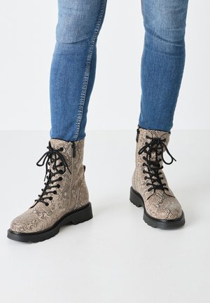 Jambes portant un jean bleu ajusté et des bottes en cuir serpent beige à lacets avec des semelles noires, debout sur un sol blanc.