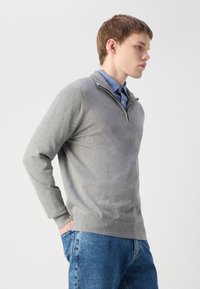Pull-over en gris clair avec un col zippé quart, des poignets et un ourlet côtelés, porté avec une chemise à col bleu clair en dessous.