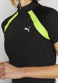 Černé tričko s krátkým rukávem a vysokým límcem, s neonově zelenými ramenními panely a malým bílým logem Puma na hrudi. Zapínání na zip u krku.